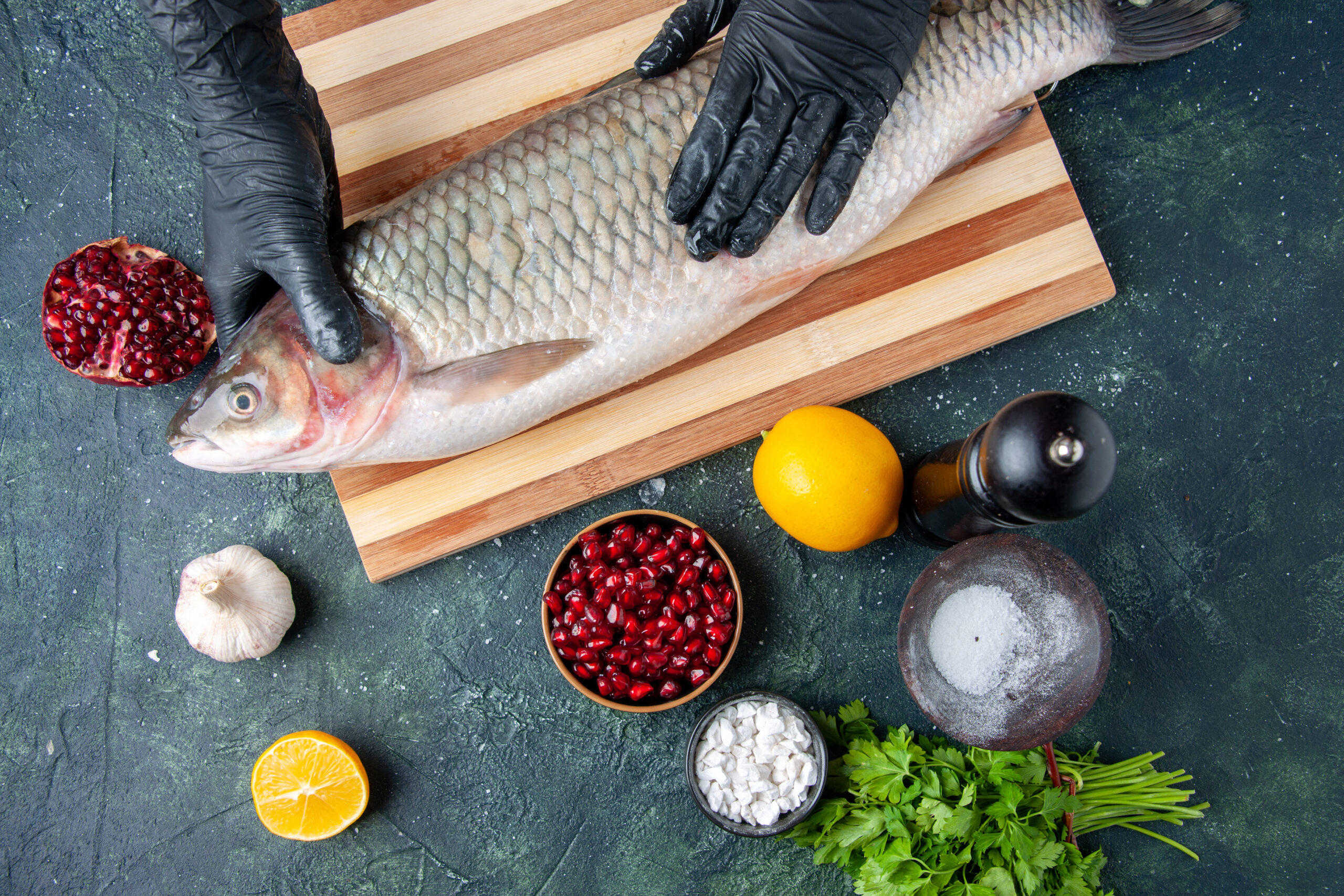 top-view-chef-with-black-gloves-holding-raw-fish-chopping-board-pepper-grinder-pomegranate-seeds-bowl-table-free-space